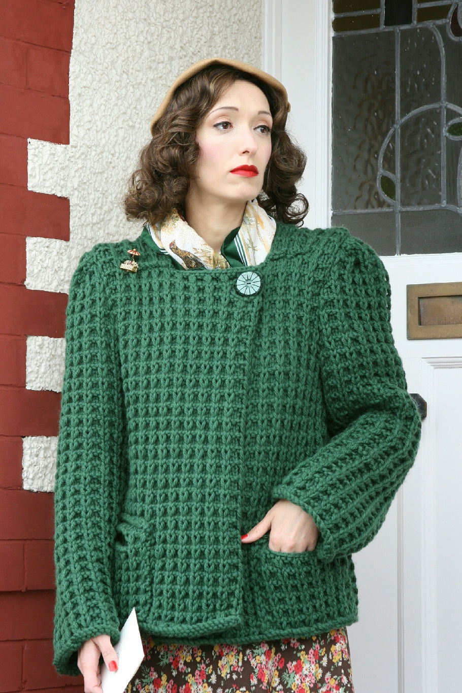 A woman stands on a doorstep, a stained glass window in the door behind her. She has curled hair and red lipstick. She is wearing a beige hat and a cream and green silk scarf around her neck. She is wearing a floral dress and over the top a green boxy knit coat with a heavy textured pattern on it. It has a large button at the neckline on one side and pockets one each side at about hip level. She is also wearing a gold brooch.