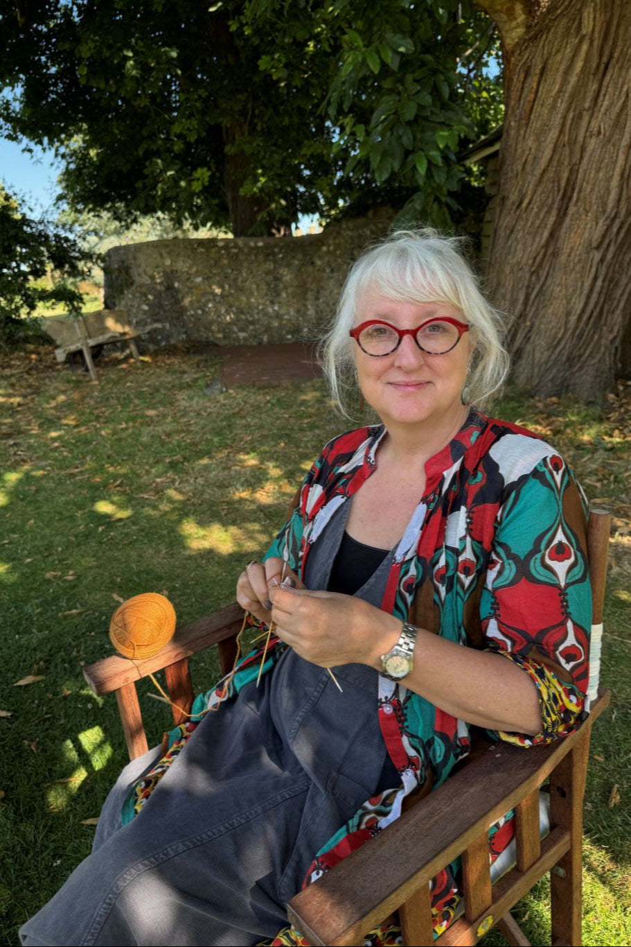 Woman with glasses and colorful scarf standing outdoors near a tree knitting socks