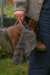A close up of a pair of worn brown leather boots being carried in one hand. Hanging from the boots are a pair of grey lacy socks.
