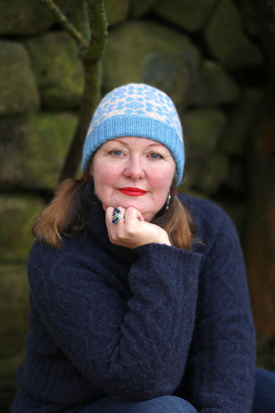 A woman with her head resting on one hand, sat down in front of an old stone wall, she is wearing a hat in a bright blue with a fair isle pattern on it.