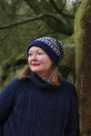 A woman looking away from the camera wearing a navy and cream fair isle hat. She is leaning against a stone wall heavy with moss.