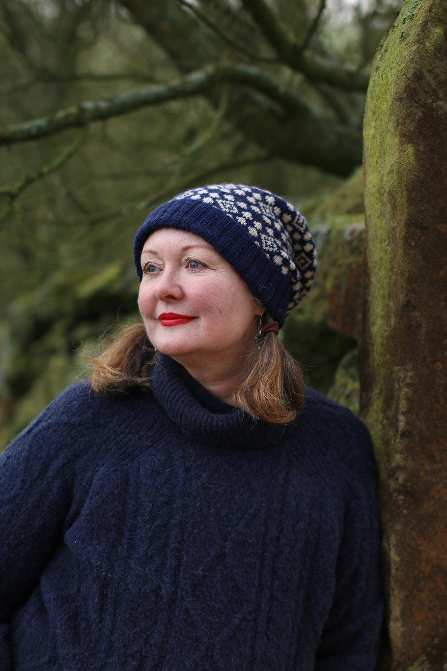 A woman looking away from the camera wearing a navy and cream fair isle hat. She is leaning against a stone wall heavy with moss.