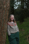 A woman leaning again a pine tree wearing a grey cardigan with a floral yoke pattern in red, pink and green