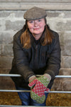 A woman resting against a metal fence wearing a wax jacket and tweed cap. She has patterned mittens on.