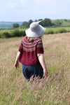 A woman in a field of long grass walking away from the camera. She is wearing a large hat with a feather in it and a short sleeved sweater with a patterned yoke.