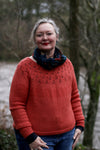 A woman wearing a red sweater with a lacy yoke standing looking at the camera and smiling. There is a river and trees behind her