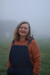 Susan standing in a misty field with hands in dungaree pockets and wearing a sweater knitted in cinnamon wool
