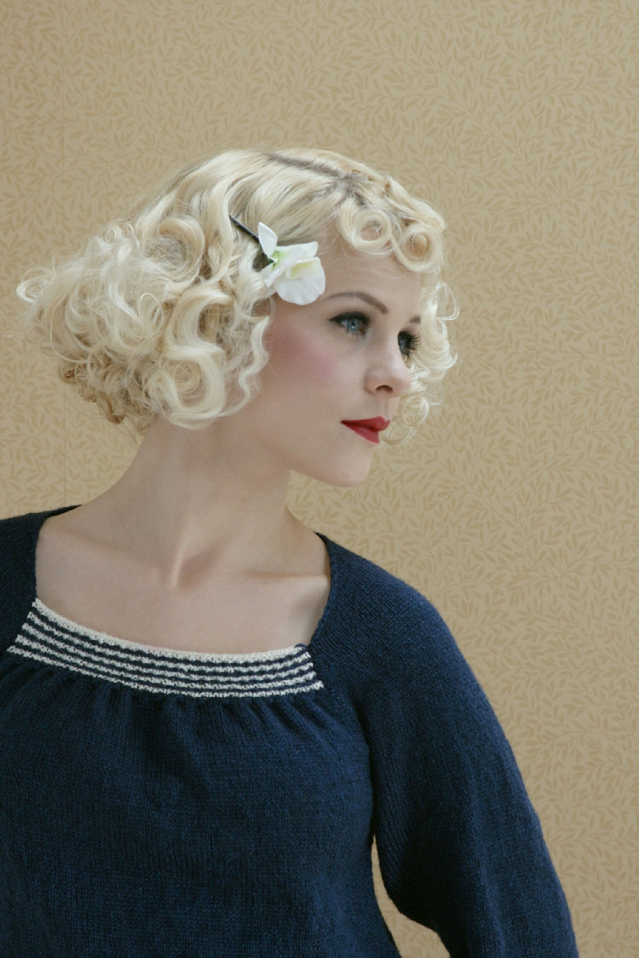 A woman curled white blonde hair stands against a pale wall. She has a white flower pinned in her hair. She looks to the side. She's wearing a navy blue knit jumper with a slightly gathered square neckline with decorative white and blue stripes across it.