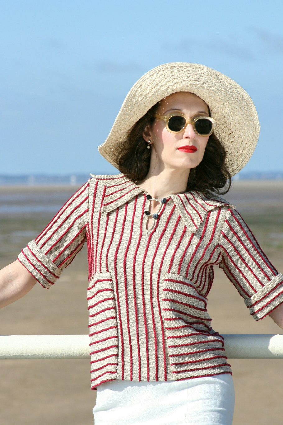 A dark haired woman stands against a railing by a beach. She wears round vintage sunglasses and a large white hat. Her top is a knit jumper in red and white stripes, both vertical and horizontal. It has a large striped collar and two buttons on an open placket neckline. The sleeves are elbow length. 