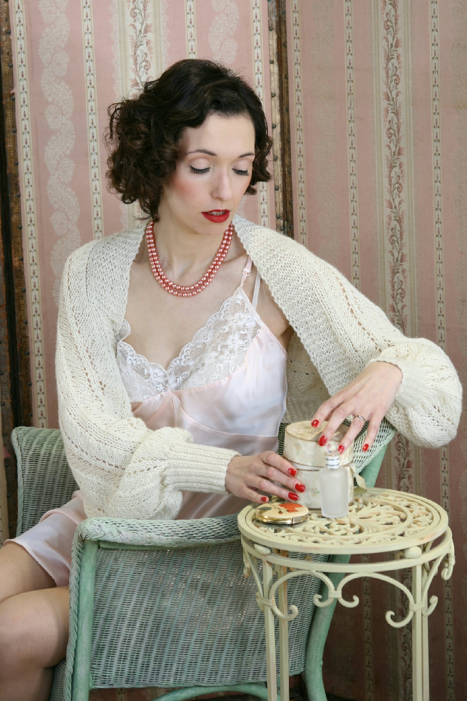 A dark woman sits by a pink papered wall, in a pale duck egg coloured rattan chair. There is a small, white, round wrought iron table to her side. There are vintage style cosmetics on the table, and she opens on of the boxes. Her dark hair is curled and she wears red lipstick and pale coral pearls. She also wears a pale pink negligee with a white lace knit shrug over the top.