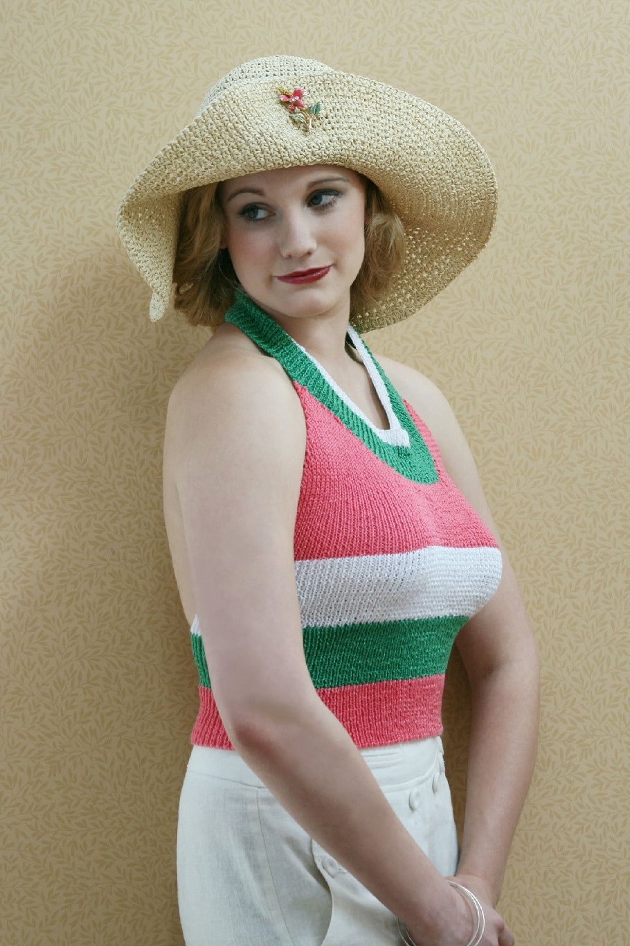 A blonde woman stands by a pale wall. She has a big natural coloured hat, the brim pinned up with a floral brooch. She stands side on to the camera and wears a knit halter neck top in white, green and pink stripes.