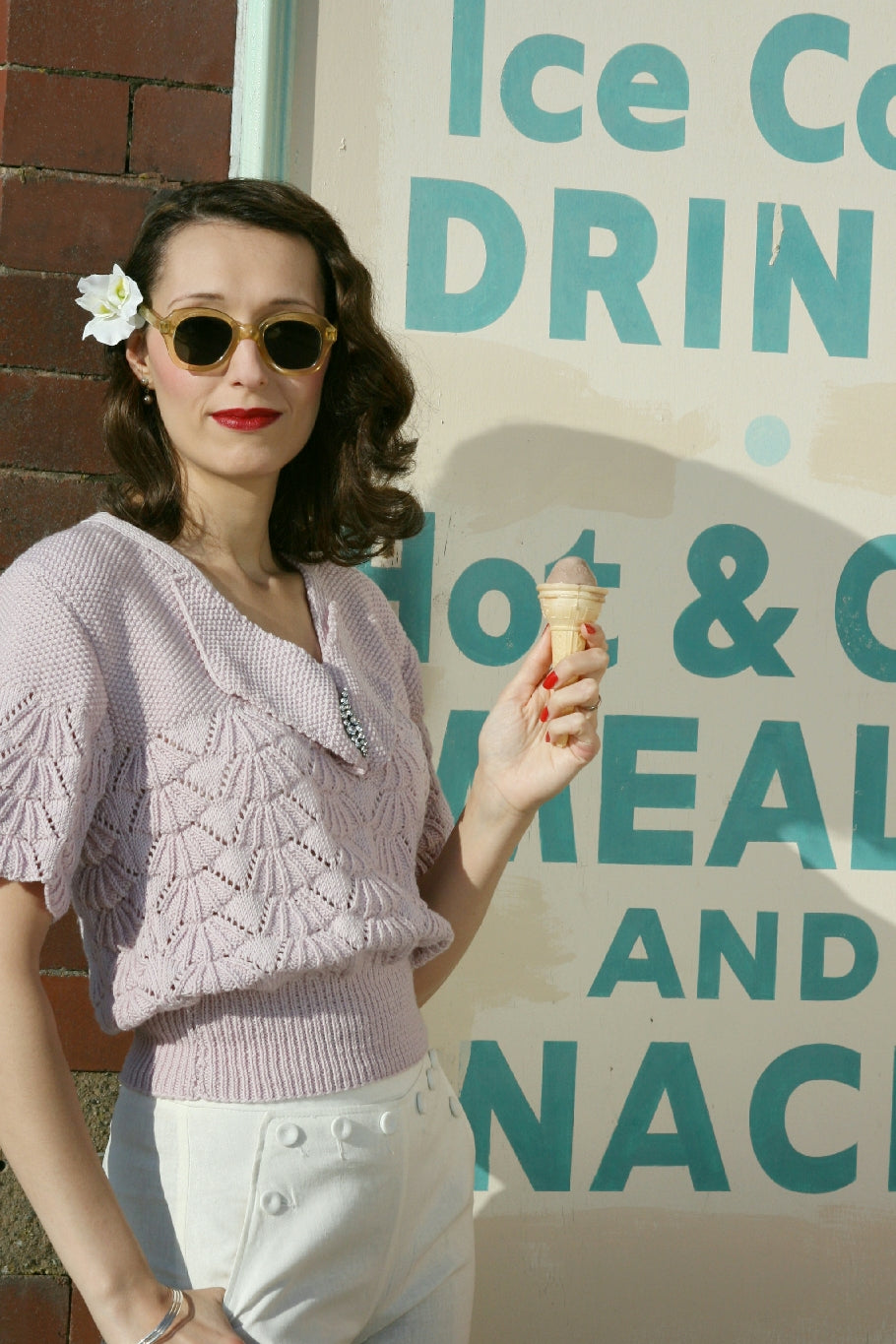 A woman stands by a sign advertising ice cream and other food and drinks. She wears sunglasses and holds a cone with icecream. Her dark hair is down in waves, with a white flower pinned behind one ear. She wears whit trousers and a vintage style knit top with a shell shaped lace pattern over the body and a large pointed collar. It has a ribbed waistband and the body of the jumper blouses out over it.