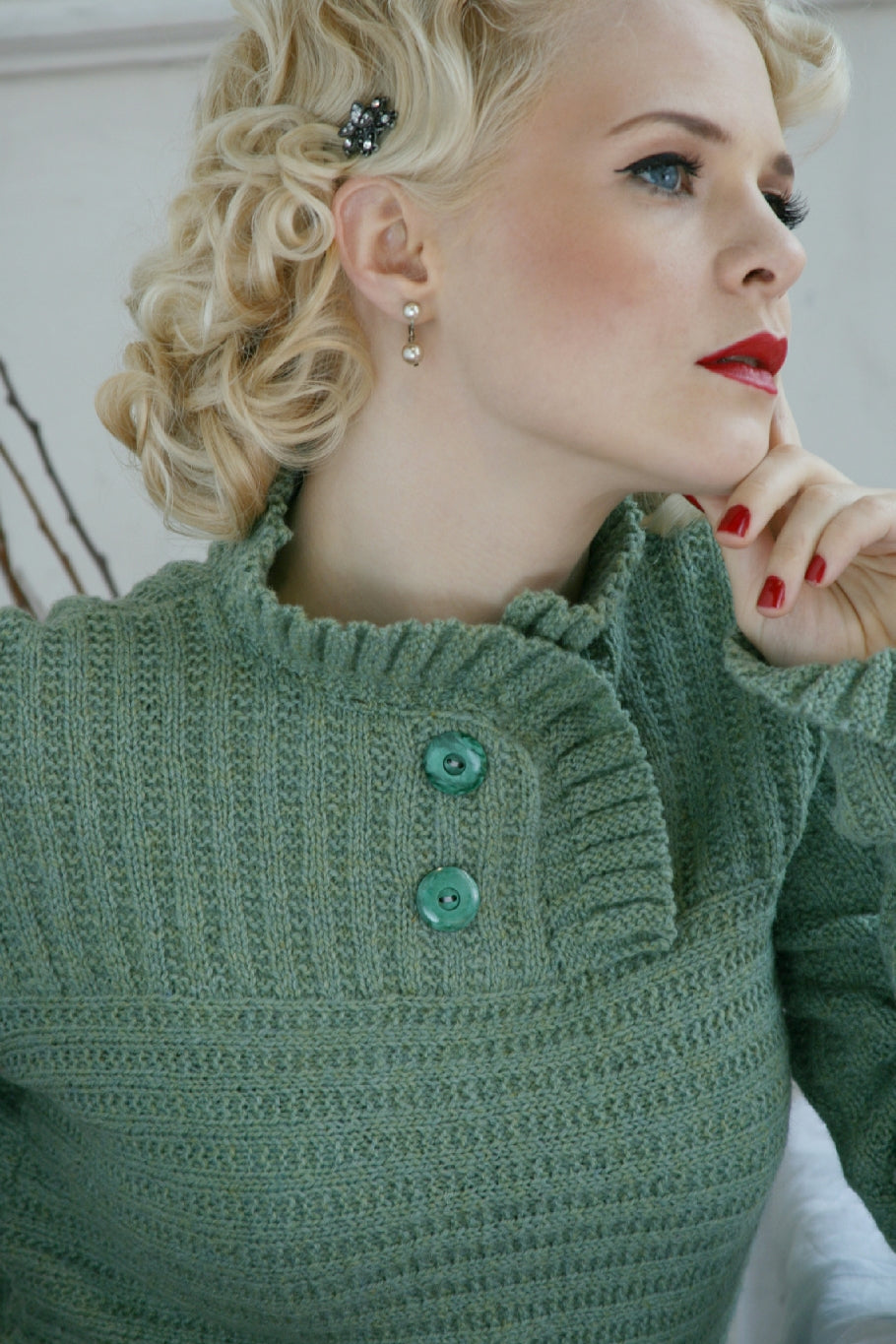 A blonde woman sits in a chair, elbow propped on the arm. She has curled hair with a vintage style clip. She wears small pearl earrings. Her fingernails are painted red, and her chin is propped on her fingers. Her jumper is a sage green knit jumper with a frilled collar. The collar comes down the placket. It has two green buttons. The pattern is a striped texture, vertical on the top of the jumper, and horizontal from the end of the placket downwards.