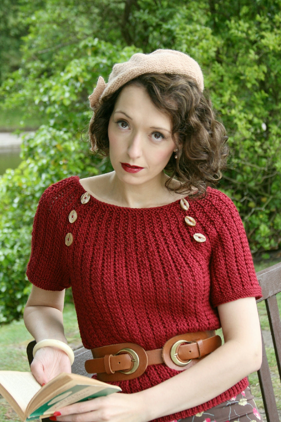 A dark haired woman leans back against a bench outdoors. She has a green Penguin Classic in her hands. She's looking up to the camera. She's wearing a natural coloured knit hat. Her top is a red textured knit jumper. It has short sleeves and buttons from the neckline to the underarm. She wears a brown leather belt around her waist.