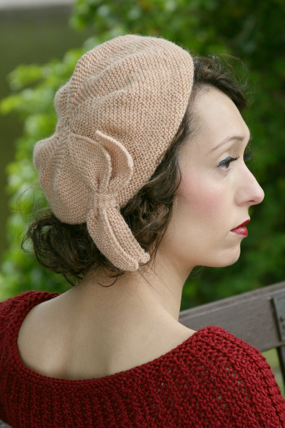 A woman sits on a bench outside. She has her back to the camera, and her face can be seen in profile. She wears a natural coloured knit hat with a knit leaf embellishment.