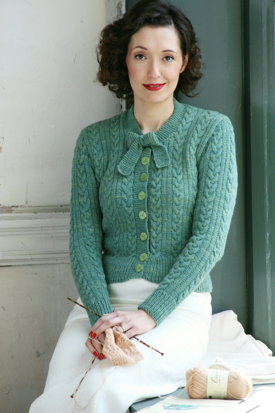 A woman with dark hair sits in a window seat. She wears a blue-green cabled cardigan with a tie at the neck and buttons down the front. She holds knitting needles and some pale pink yarn.