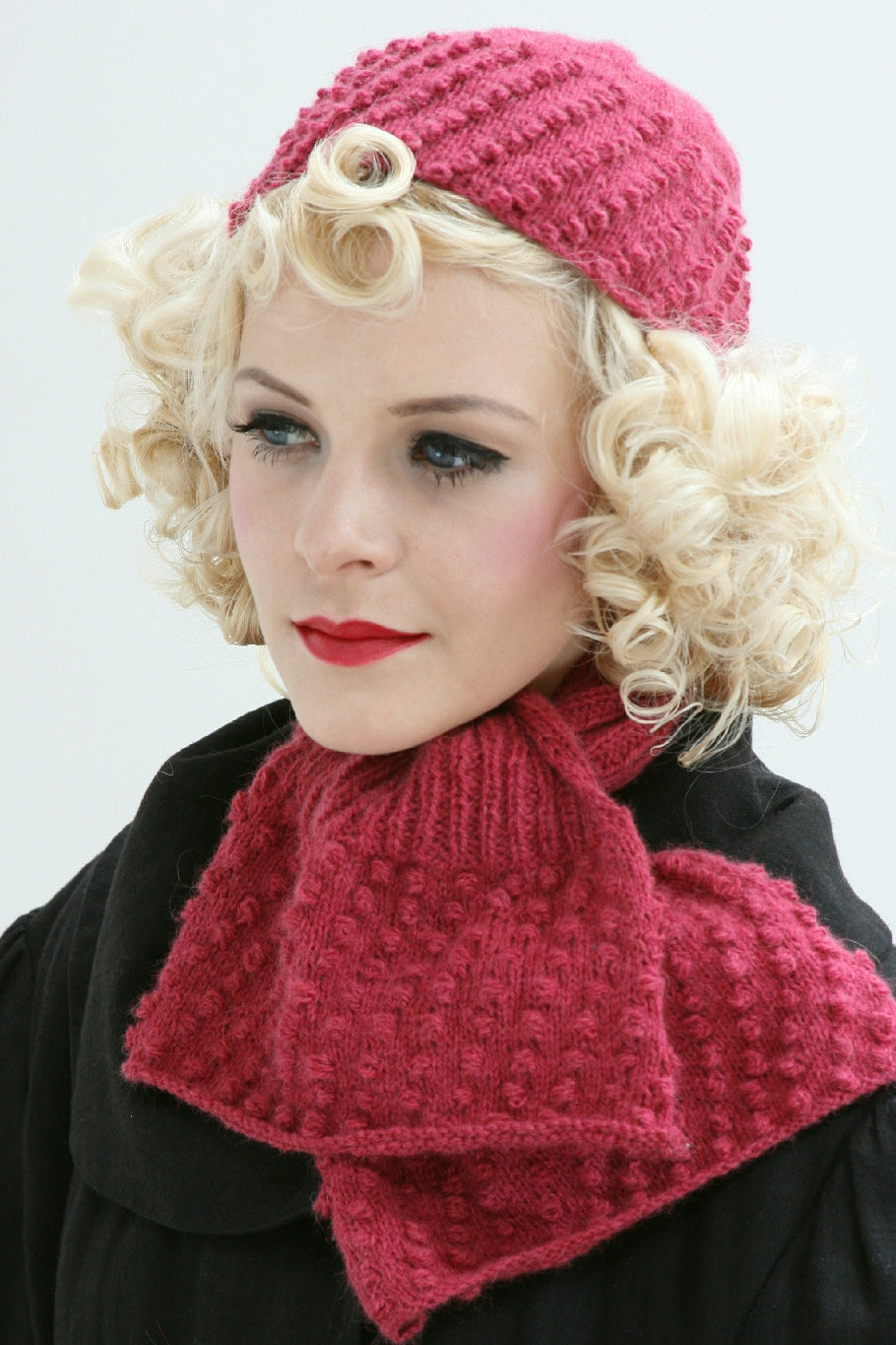 A woman with blonde curly hair is wearing a raspberry coloured cap and scarf with a rib and bobble pattern. She has a black coat and stands by a white background.