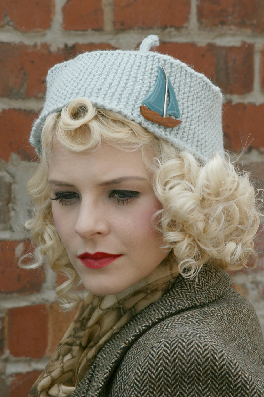 A blonde woman with curly hair is in front of a brick wall. She wears a tweed coat and a patterned silk scarf around her neck. She wears a pale blue knit fez with a boat pin decorating the side.  It has a stem coming up from the centre top.