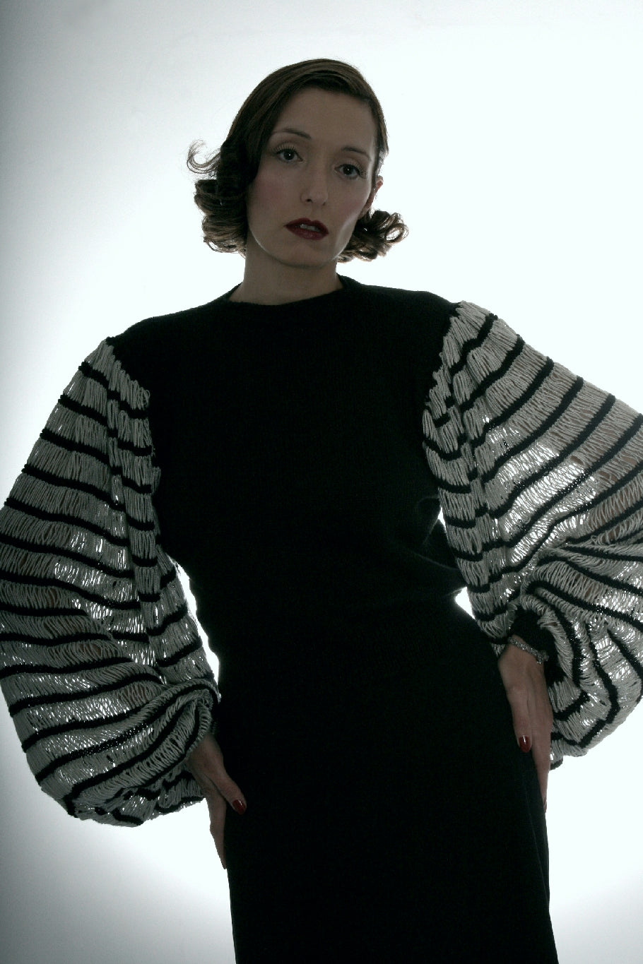 A dark haired woman is standing, hands on hips, with vintage styled hair and make up. She wears a black knit jumper with very big floppy sleeves, striped in black and white. She is in front of a brightly lit background.