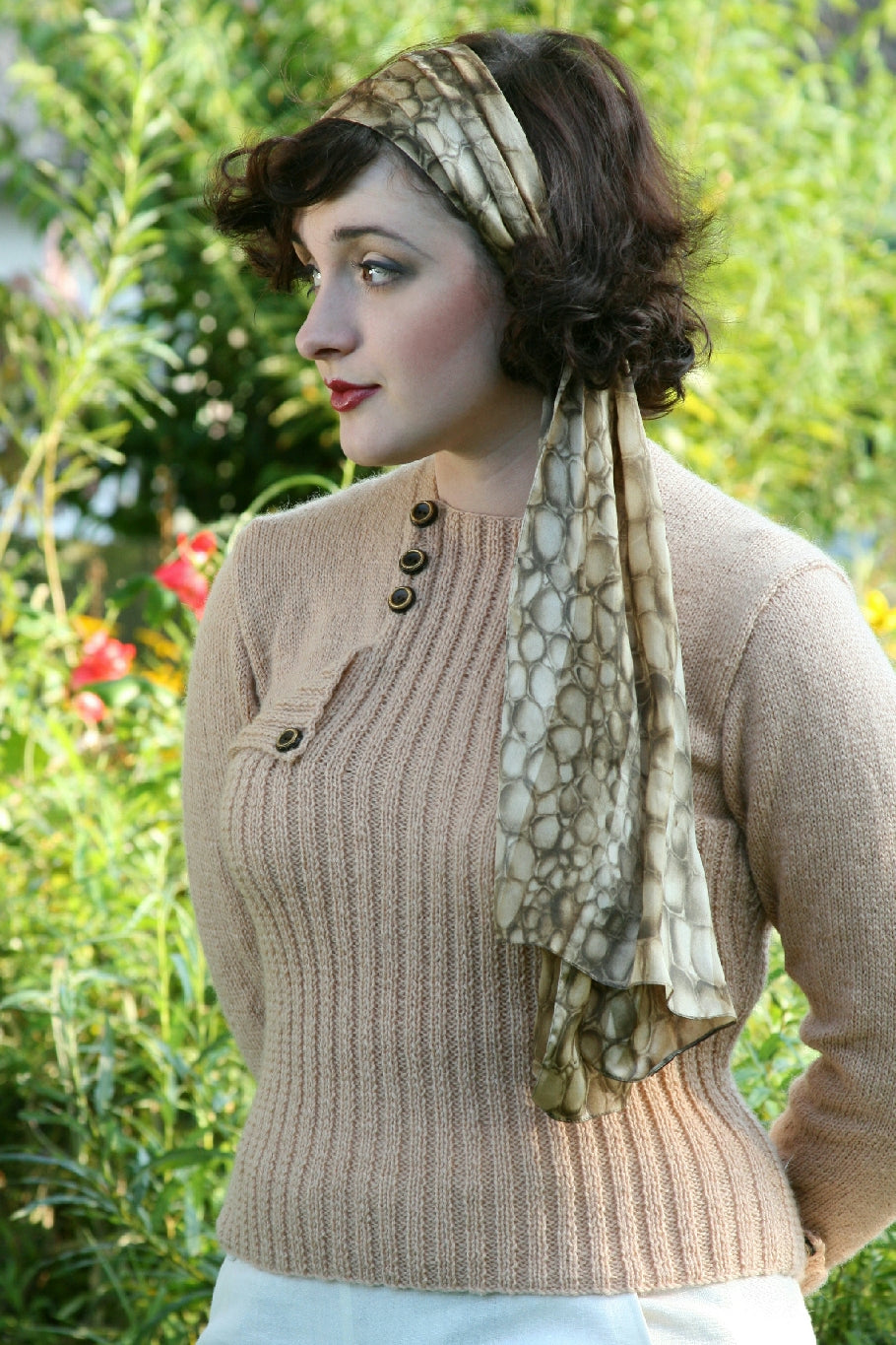 A woman with dark hair tied in a silk scarf is standing outside. The scarf has an organic circular pattern and hangs down over one shoulder. She wears a natural knit jumper with ribbing down the front. The shoulders and sleeves are stocking stitch. There are 3 accent buttons where the shoulder and neck met on one side, and another over a pocket.