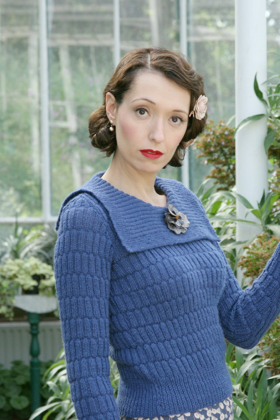 A woman stands, surrounded by plants. She's standing side on to the camera, her face towards it. She has vintage styled hair, with a flower pinned on one side. She also has small peal drop earrings. She's wearing a blue knit jumper with a lozenge pattern over the body and sleeves. It has a square sailor style collar and is decorated with a flower brooch.