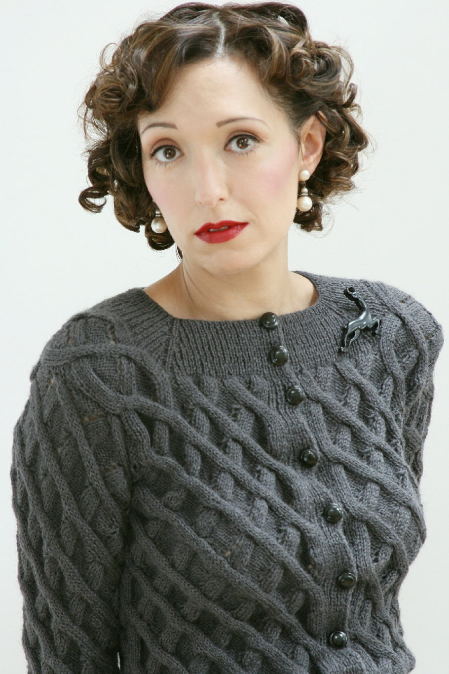 A woman stands by a white background. She has dark curled hair, red lipstick and pearl drop earrings. She is wearing a dark grey cardigan. It has a heavily cabled design, and black buttons up to the neck. She also wears a dark brooch shaped like a monkey.