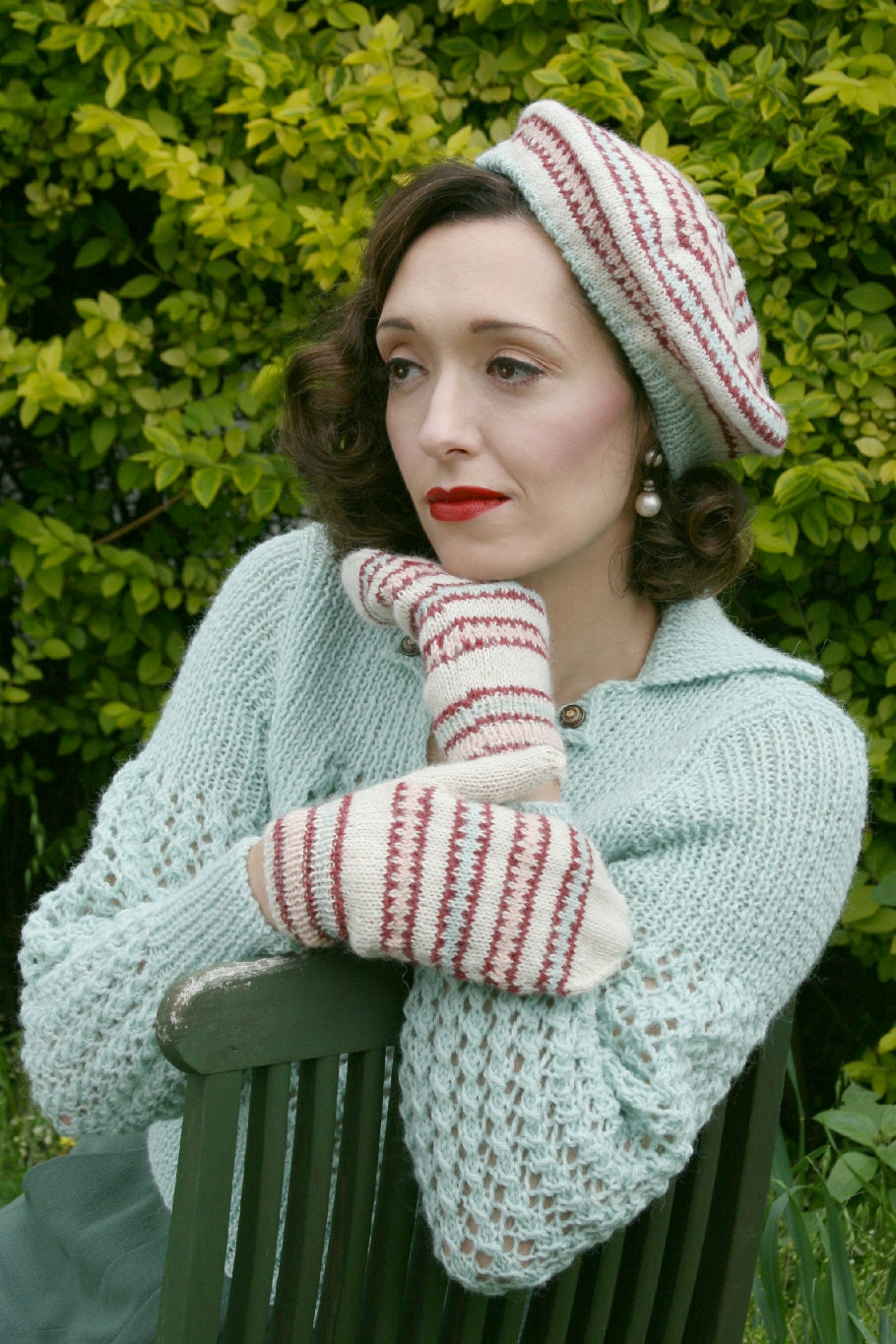  A dark haired woman sits on a green garden chair against a backdrop of leaves. She has a Fair Isle hat on top of her dark curls, and pear drop earrings below. She's wearing a light blue knit jumper with lace sleeves. One hand is under her chin and the other rests on her arm. She is wearing Fair Isle mittens in the same natural, blue and red pattern as her hat.