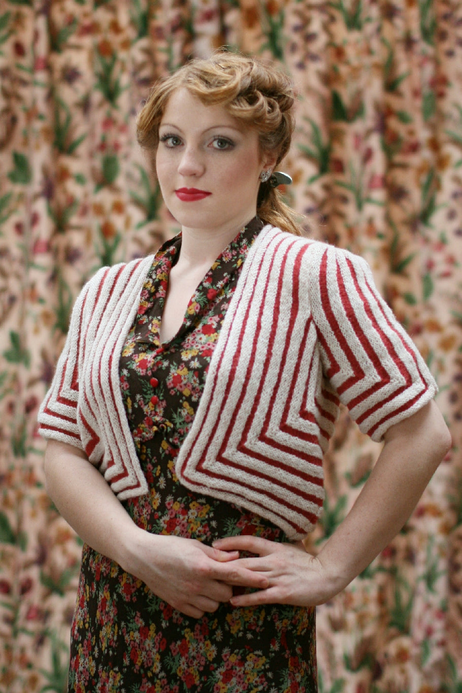 A woman with vintage styled hair stands in front of a floral curtain. She wears a black and coloured floral dress. Over the top she wears a red and white knit cardigan in a square shape with mitred corners on the cardigan fronts and on the sleeves.