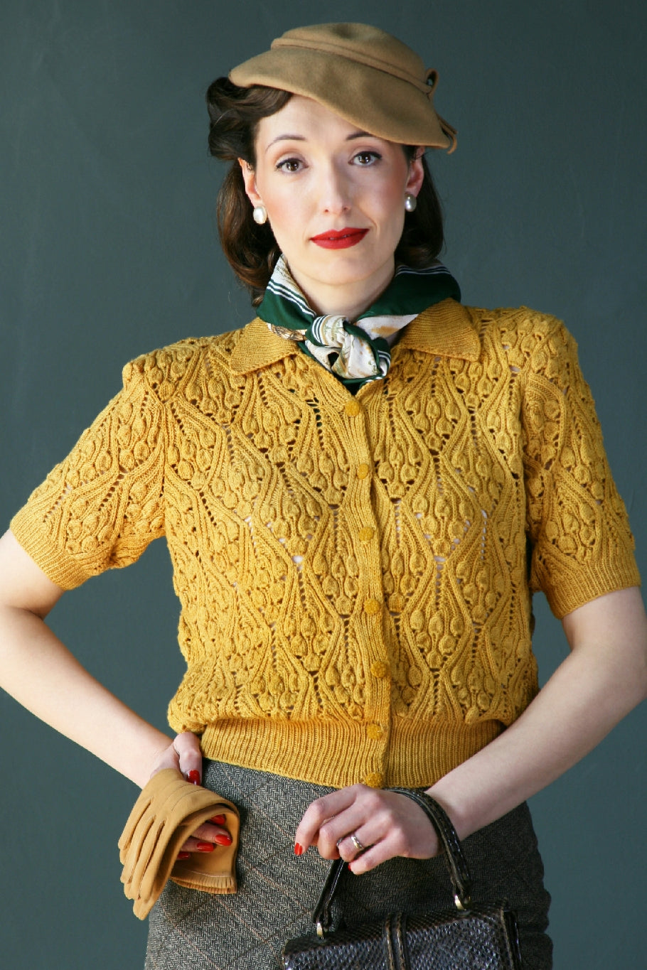 A dark haired woman stands in front of a blue-grey background. She has vintage styled hair and hat as well as red lipstick. She has a green and cream silk scarf tied at her neck. She's wearing a yellow knit lace cardigan with a pointed collar and sleeves that fall just above the elbow. She's also holding a mustard pair of leather gloves in one hand and a black handbag in the other.