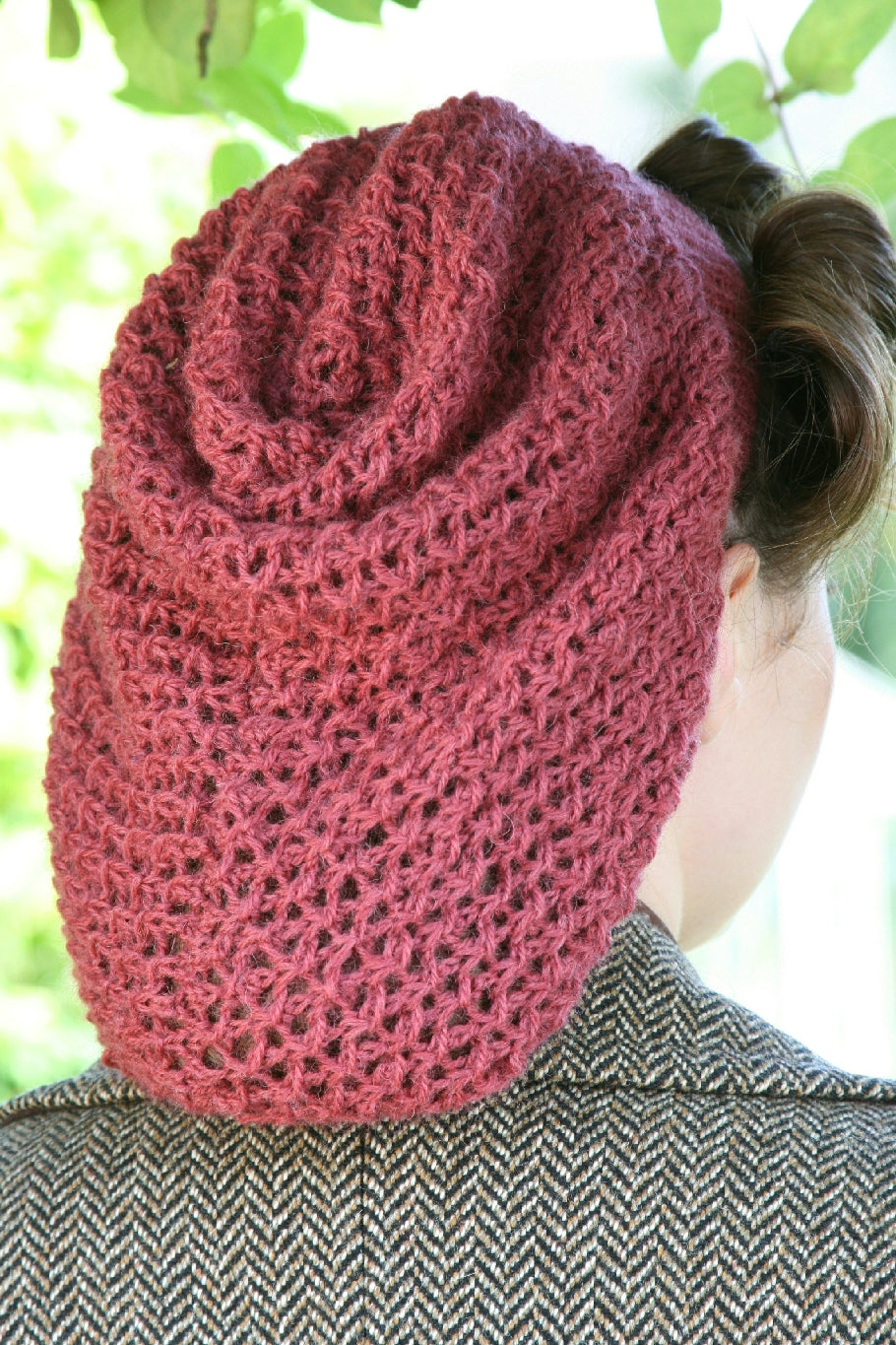 A woman is standing outside. She is facing away from the camera. She is wearing a brown tweed coat and her brown hair is in 40s style rolls. She's wearing a dusky pink snood in a knit lace pattern. The snood comes down to the bottom of the coat's collar.