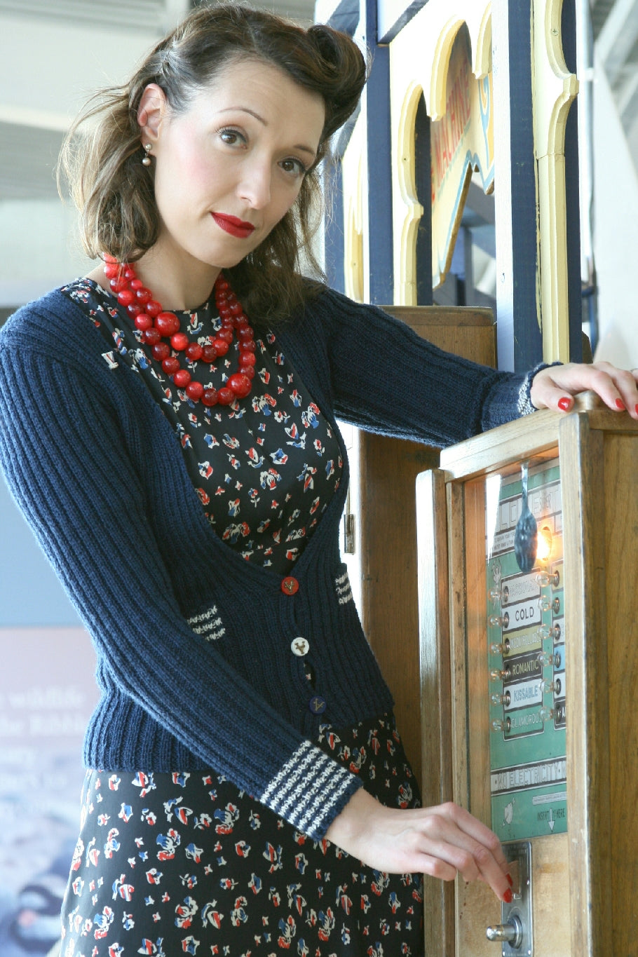 A woman with dark 1940s styled hair stands by a vintage vending machine. One hand is resting on top of the machine and the the other is putting a coin into the slot. She's wearing a black and floral dress and two strands of large red beads. She's wearing a navy blue knit cardigan with a deep v. There are white stripes at the top of the pockets and at the cuffs of the sleeves. It has three buttons, a red one, a white one and a blue one.