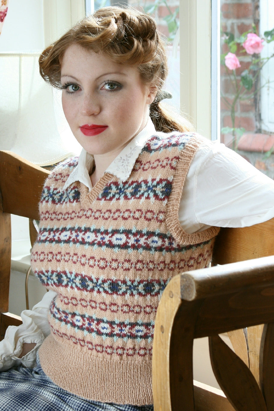 A woman with 1940s styled dark blonde hair sits on a wooden bench by a window. She wears a white blouse and a Fair Isle knit vest over the top. The vest is in apricot, blue, yellow, red and white.