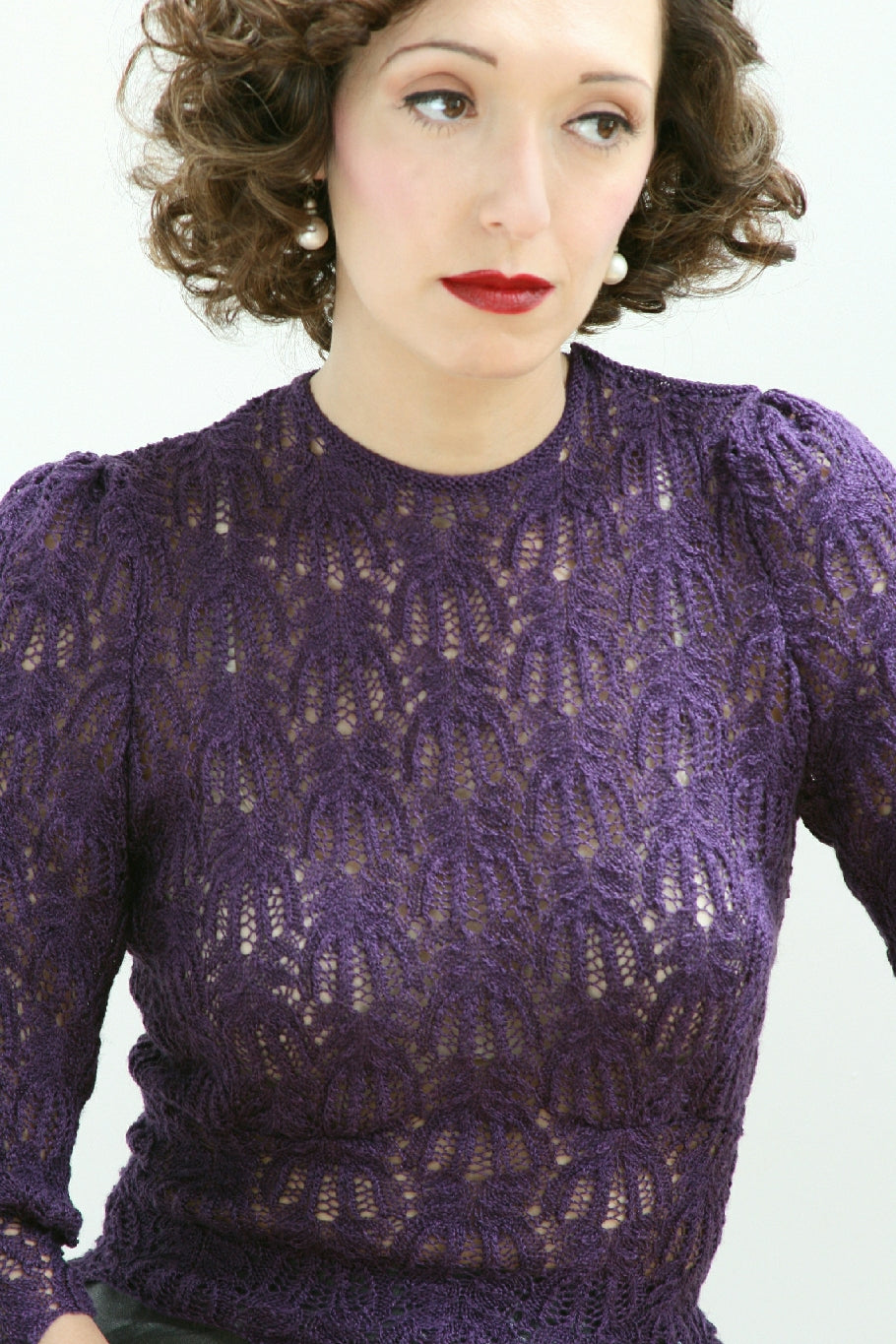A woman is pictured with a white background behind her. She has dark curly hair, red lipstick and pearl drop earrings. She is wearing a high necked purple lace knit jumper with slightly puffled long sleeves.