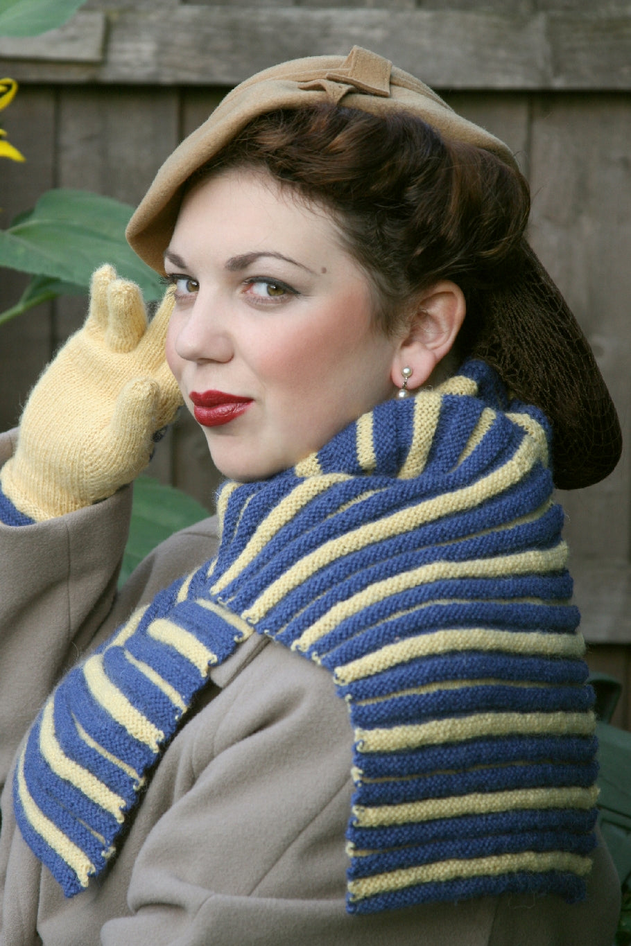 A woman with dark hair in a net snood, with a beige vintage style hat is sitting outside on a bench by a wooden fence. She is sitting in profile, with her head slightly turned towards the camera. She is wearing a beige coat and pale yellow knit gloves with a blue and white striped knit scarf. The scarf has a ridged design.