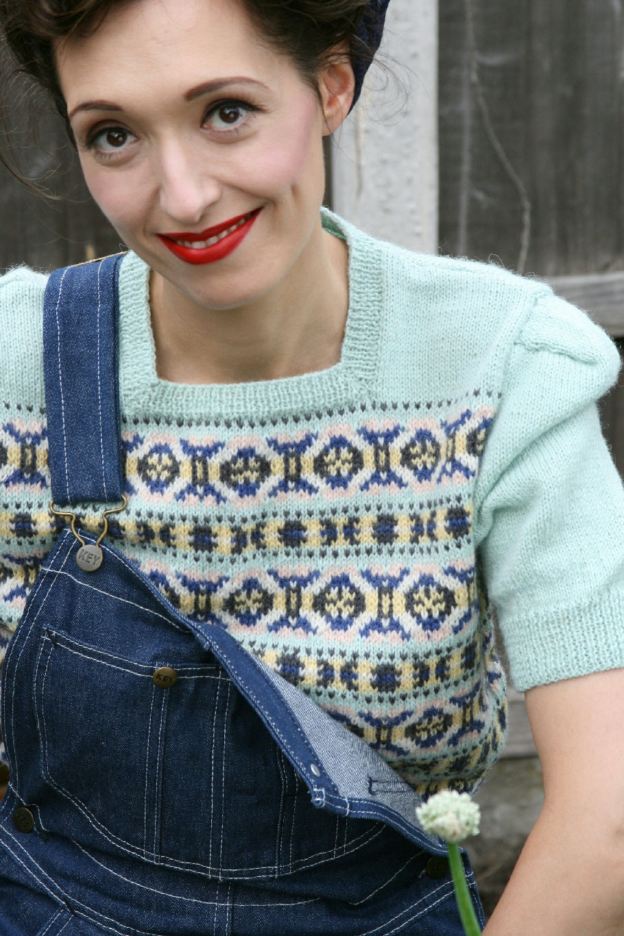 A dark haired woman is outside. She is wearing dark blue denim dungarees, with one clasp undone. Underneath she wears a pale blue knit jumper with a square neckline and short puffed sleeves. There is a Fair Isle design in blue, black and white on the body of the jumper.