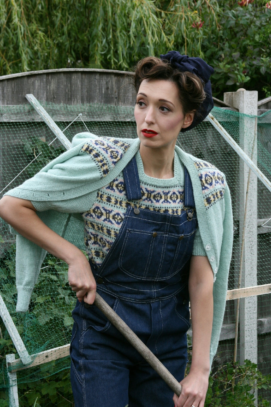 A woman is outside, wearing her hair tied up in a navy blue scarf. She's wearing dark denim dungarees over a Fair Isle short sleeved jumper, and there is a matching cardigan draped over her shoulders. The jumper and cardigan are pale blue with blue, red, yellow and white Fair Isle patterns on them. She is holding the handle of some garden equipment.