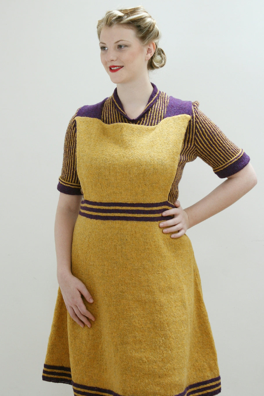 A blonde woman, her hair styled in 1940s style, stands against a white background, one hand on her hip. She is wearing a yellow and purple knit pinafore dress. The straps are purple and there are three purple strips at the waist and the hem of the dress. She is wearing a coordinating knit jumper in yellow and purple stripes. There is a narrow collar, and purple trim at the cuffs of the elbow sleeves.