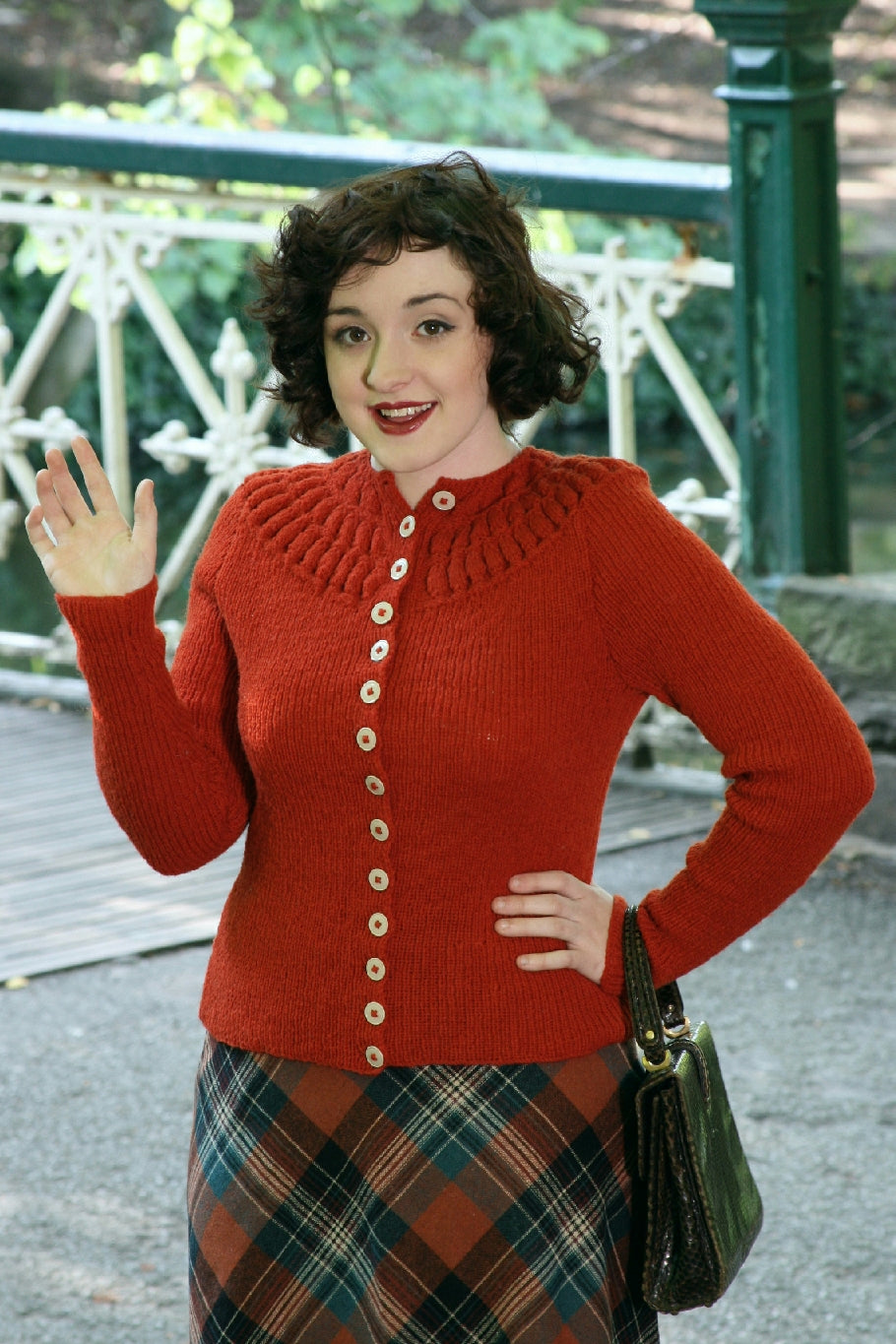 A dark haired woman is standing outside, some green and white wrought iron railings behind her. She is wearing a tartan skirt and a red knit cardigan. The cardigan has a high neck with a bubble stitch on the yoke. She has one hand on her hip, and a handbag held on her wrist. The other hand is held up as if to wave.