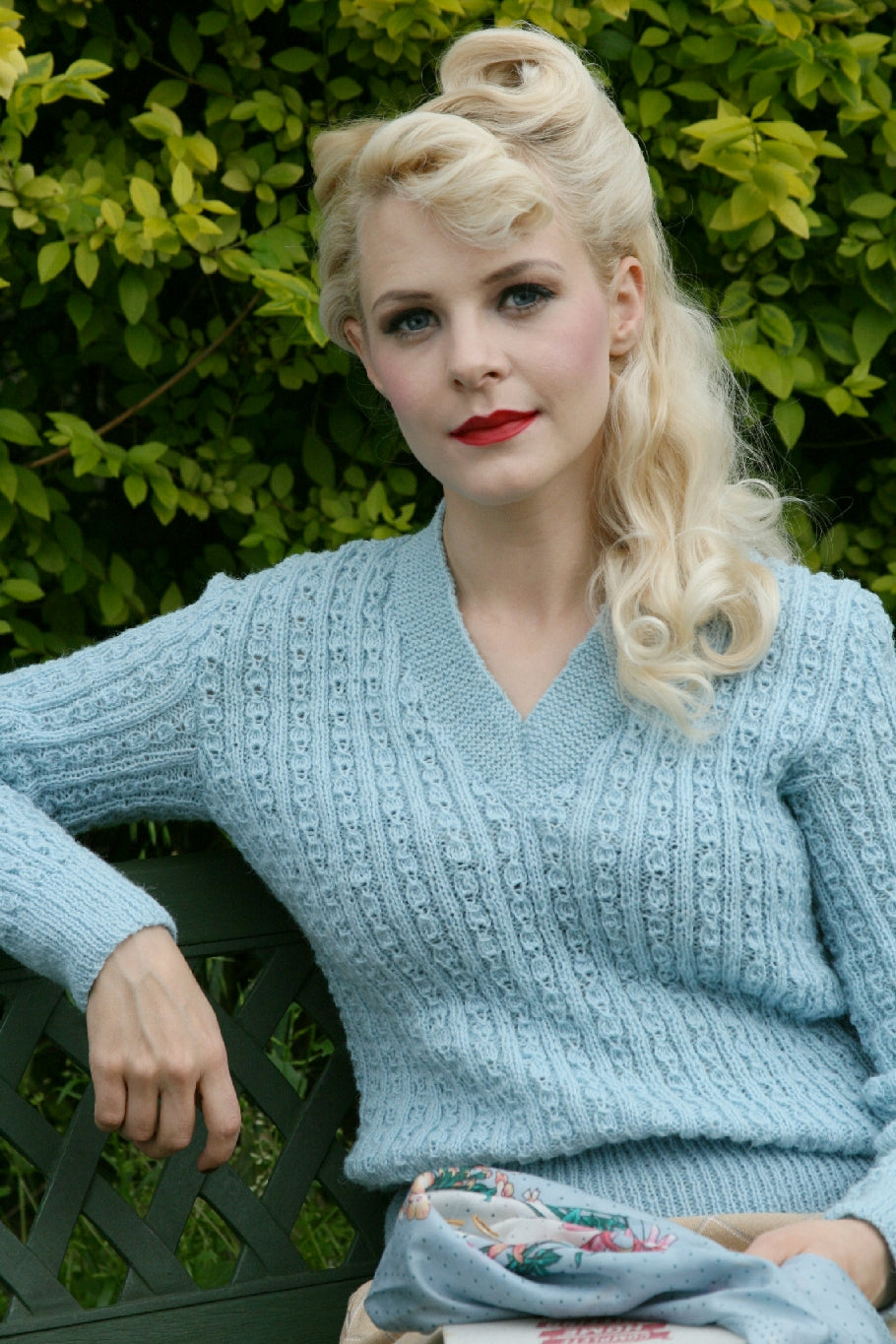 A blonde woman sits by a hedge on a green wooden bench. She has her hair down but in 1940s rolls on top of her head. She is wearing a pale blue v-neck jumper with a ribbed and lace pattern across it. The jumper has long sleeves.