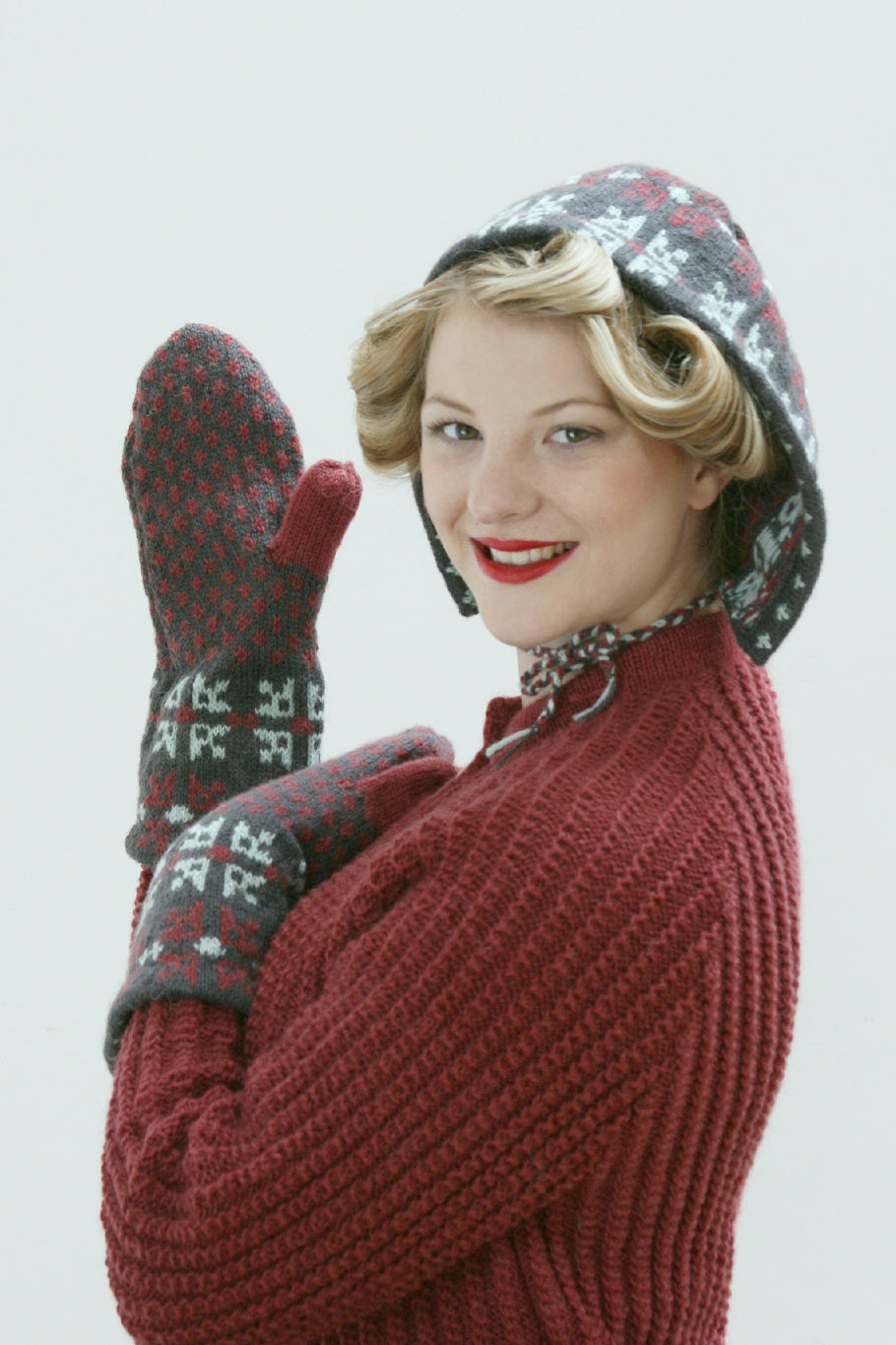 A blonde woman stands against a white background. She is wearing a red knit jumper, and a knit bonnet and mittens. They are both grey, red and white with a Fair Isle pattern. There is a knit tie at the neck of the bonnet. She is standing in profile and has one hand up showing the palm of one of the mittens.