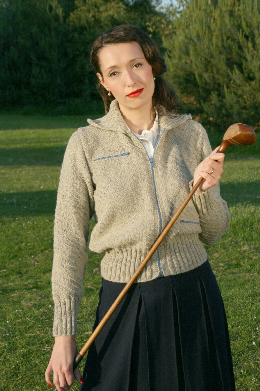 A woman stands outside. She has dark curled hair and is holding a wooden golf club. She is wearing a dark pleated skirt and a cream knit jacket with a blue zip and blue accents on the pockets.