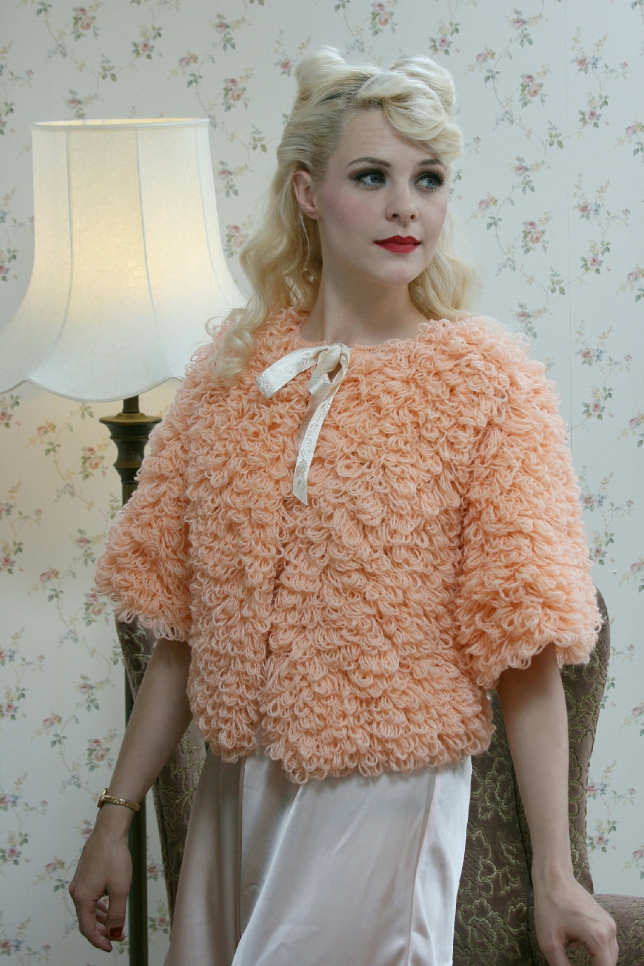 A blonde woman with vintage styled hair and makeup stands in front of an arm chair and a standard lamp. There is a delicate floral wallpaper behind her. She is wearing a peach knit bedjacket with a looped textured stitch and a satin ribbon bow at the neckline. The sleeves are about elbow length. She wears the bedjacket over a satin nightdress.