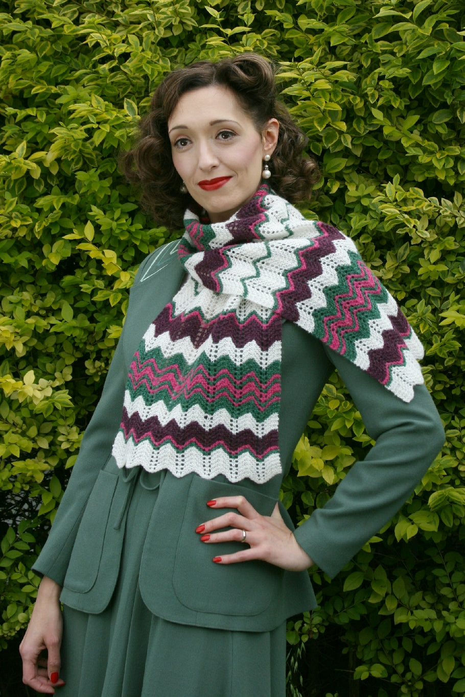 A dark haired woman stands in front of a hedge. She has vintage styled hair and makeup and wears a sage green skirt suit, also in a vintage style. Around her neck is a wide scarf in a feather stitch with chevron stripes of white, green, brown and pink.