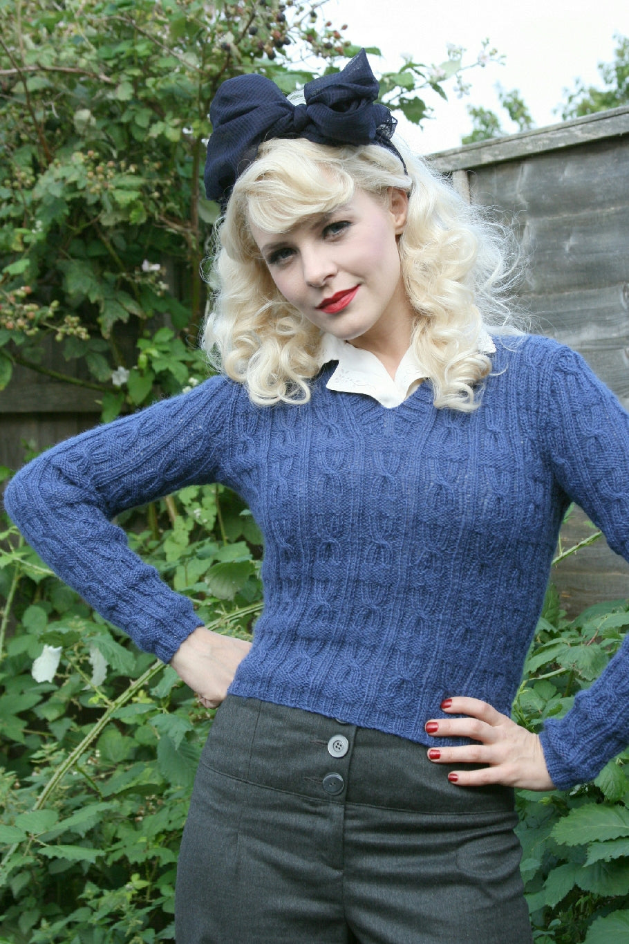 A blonde woman stands outside in a garden. She has a large black scarf tied in a bow on her head. She wears high waisted trousers and a white shirt with a blue cableknit jumper. She is standing with her hands on her hips while smiling slightly at the camera.