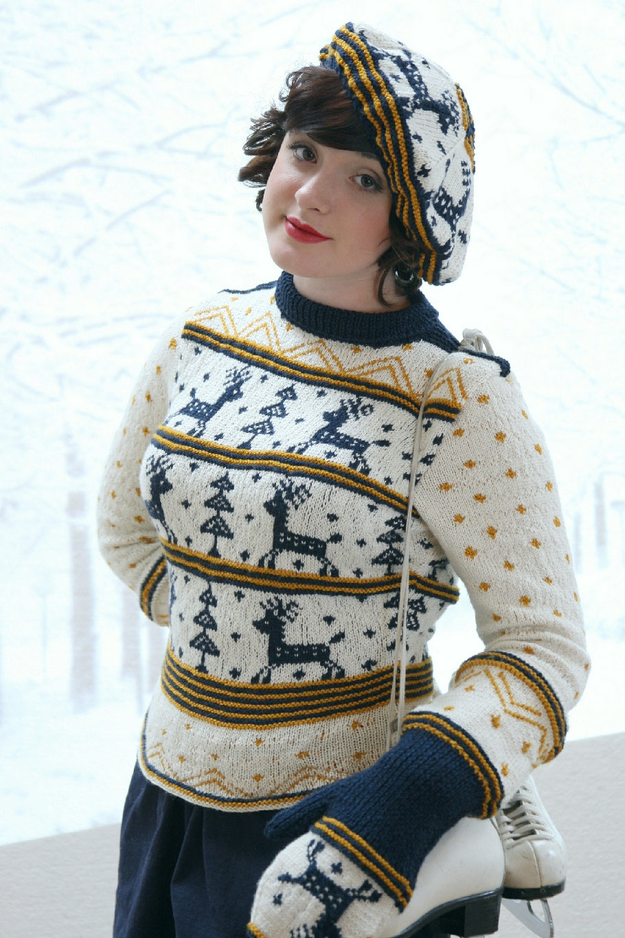 A dark haired woman stands by a wintery background. She has one hand on her hip, and head tilted as she looks to the camera. She has a pair of ice skates slung over one shoulder. She is wearing a mating jumper, hat and mittens set.  They have a Scandinavian Christmas feel. It has reindeer and pine or Christmas trees in navy blue on a white background with other decoration and stripes in golden yellow and the same navy blue.