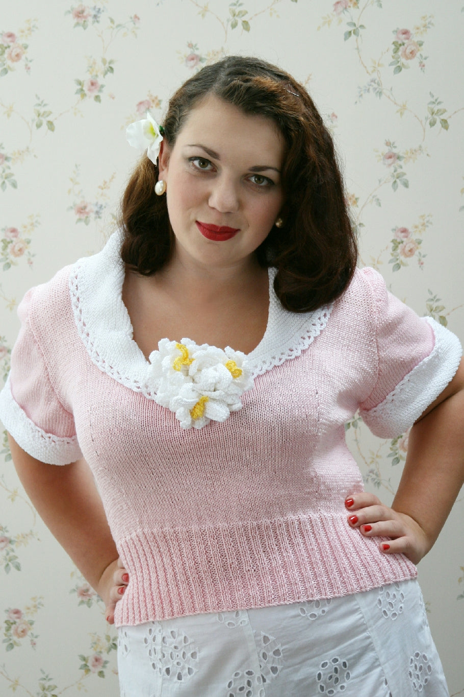 A dark haired woman with a white flower behind her ear, red lipstick and pearl earrings faces the camera. Her hands are on her hips. She is wearing a short sleeved knit jumper in pale pink. It has white lace edged collar and cuffs, and flowers at the centre of the v-neckline. 