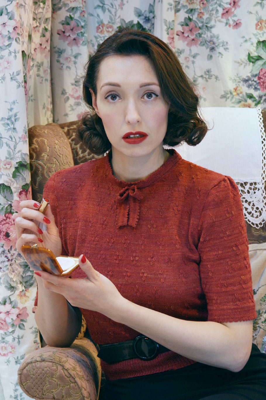A dark haired woman sits in a vintage armchair with antimacassar. A floral curtain is behind her. She faces the camera and holds lipstick in one hand and a vintage makeup compact in the other. She looks as though she has just applied her lipstick. She is wearing a dark rust knit top with a gathered bow at the neckline.