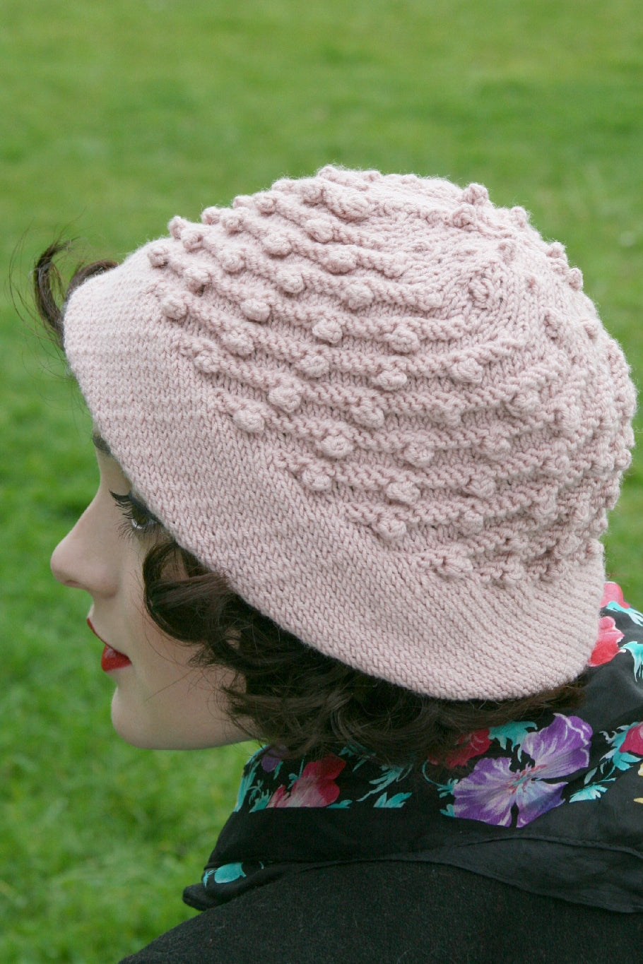A dark haired woman sits outside. She is wearing a black coat and a floral scarf. On her head is a natural cream coloured hat with a plain brim and a bobbled pattern on the main body of the hat.