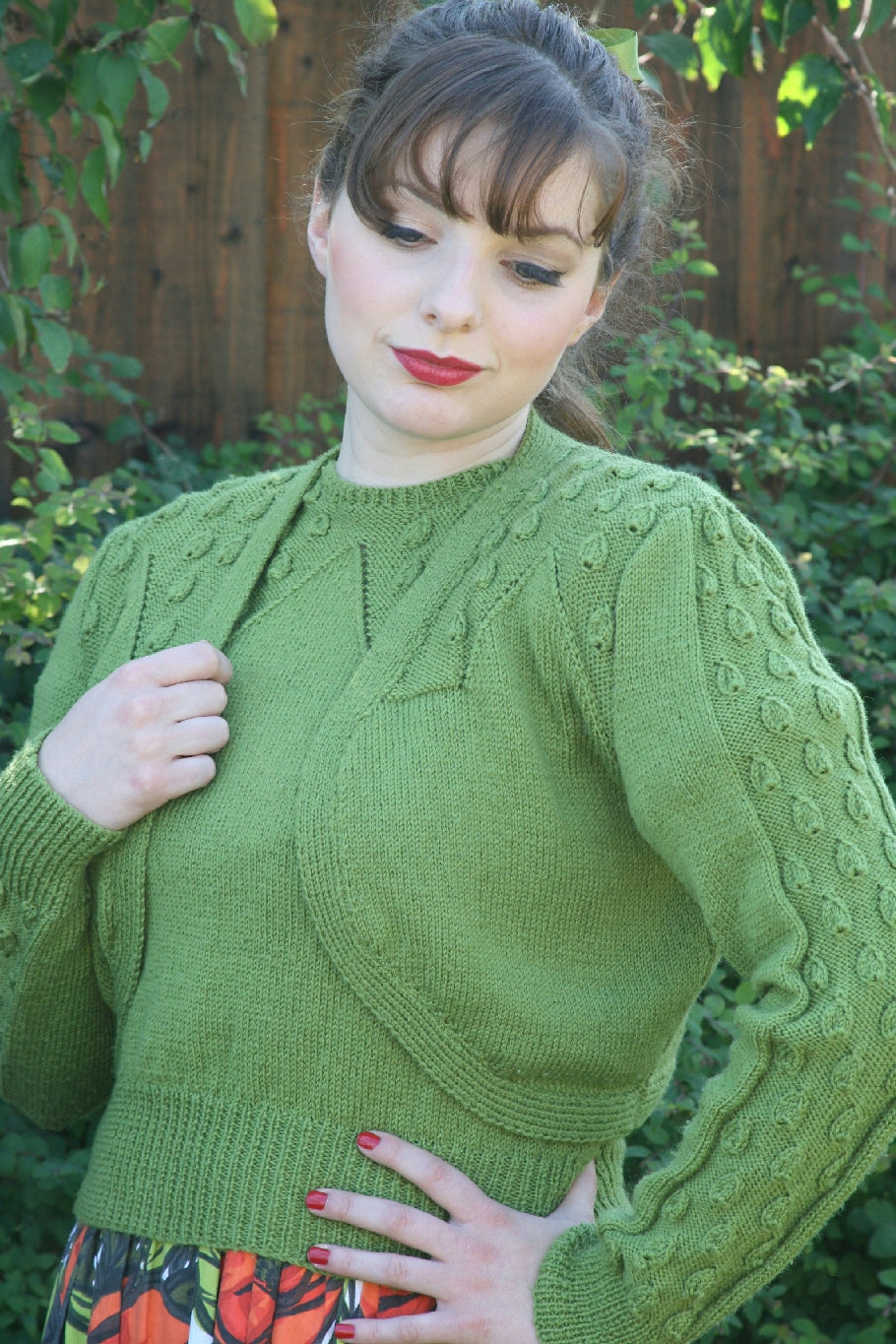 A dark haired woman stands outside with trees and greenery behind her. She has her hair up in a ponytail and a long fringe over her forehead. She is wearing a matching knit top and bolero in green yarn. The shoulder and sleeves of the bolero feature a droplet pattern. She is also wearing a floral skirt. One hand is on her hip and the other holds the edge of the bolero on one side.
