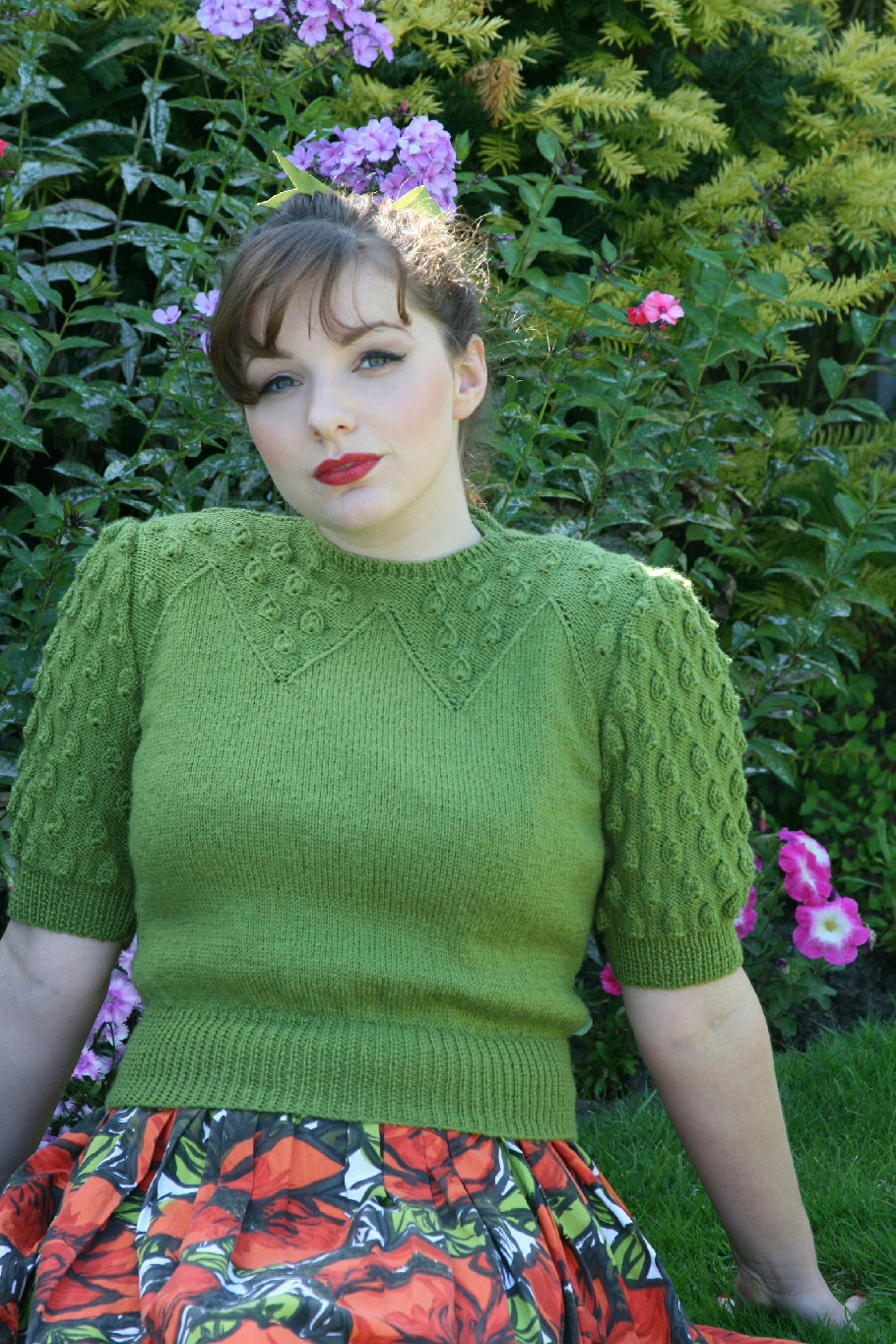 A dark haired woman sits on the grass outside, leaning back on her hands. She has dark hair in a ponytail and a long fringe. She is wearing a green knit top with elbow length sleeves and a floral skirt. The top has a decorative droplet pattern on the sleeves and part of the neckline.